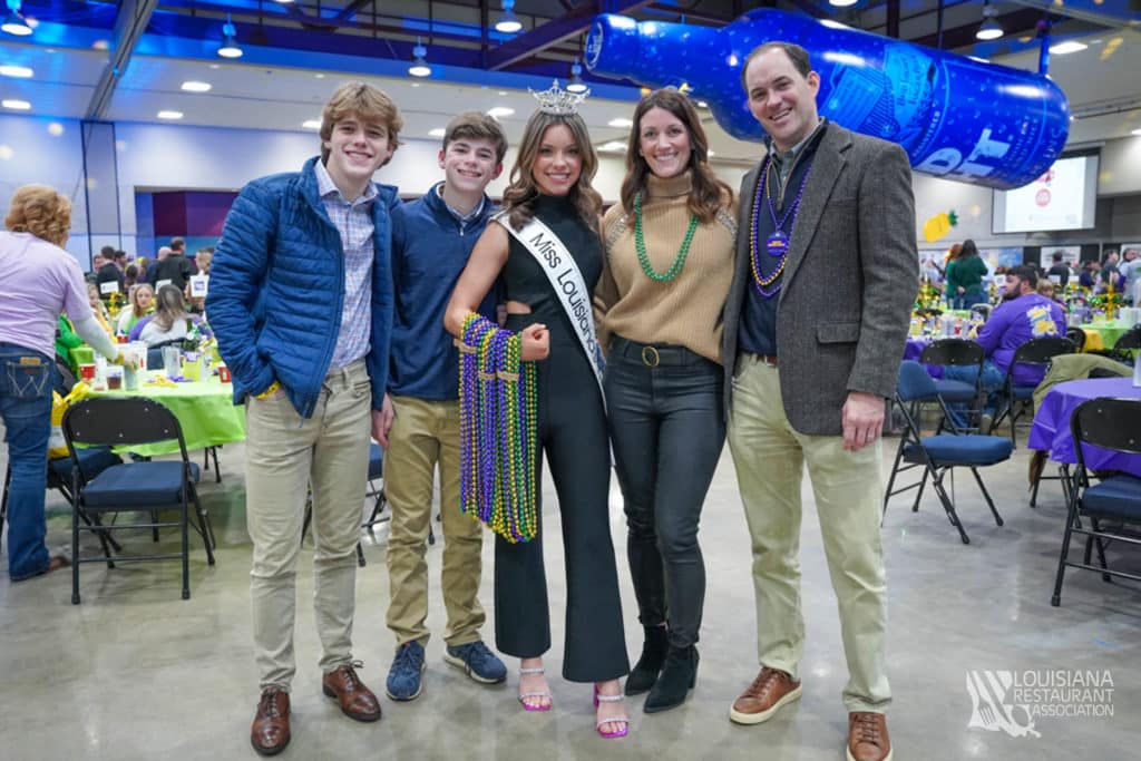 a group photo of people, one in a white dress with a sash, in an indoor setting.