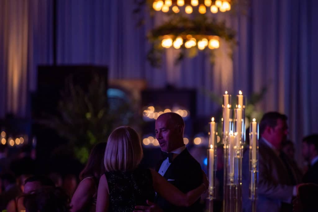 A couple in silhouette converse under the soft glow of a grand chandelier, with elegant candlesticks in the foreground, at a dimly lit social event.