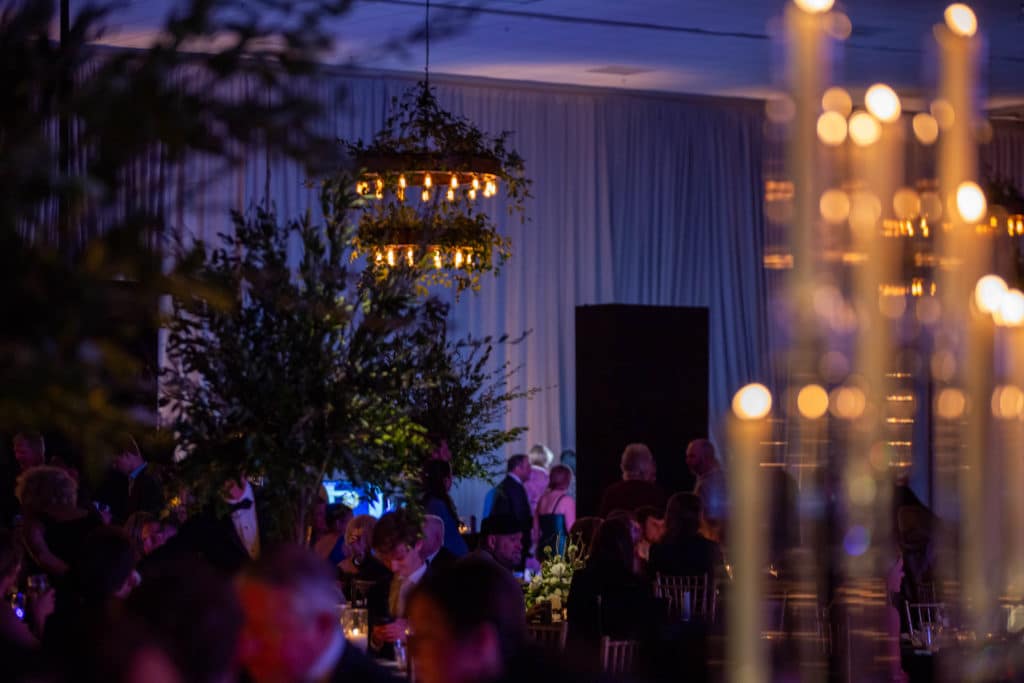 Low-lit event venue with blue ambient lighting, guests mingling, and a chandelier visible in the background, creating an intimate atmosphere.